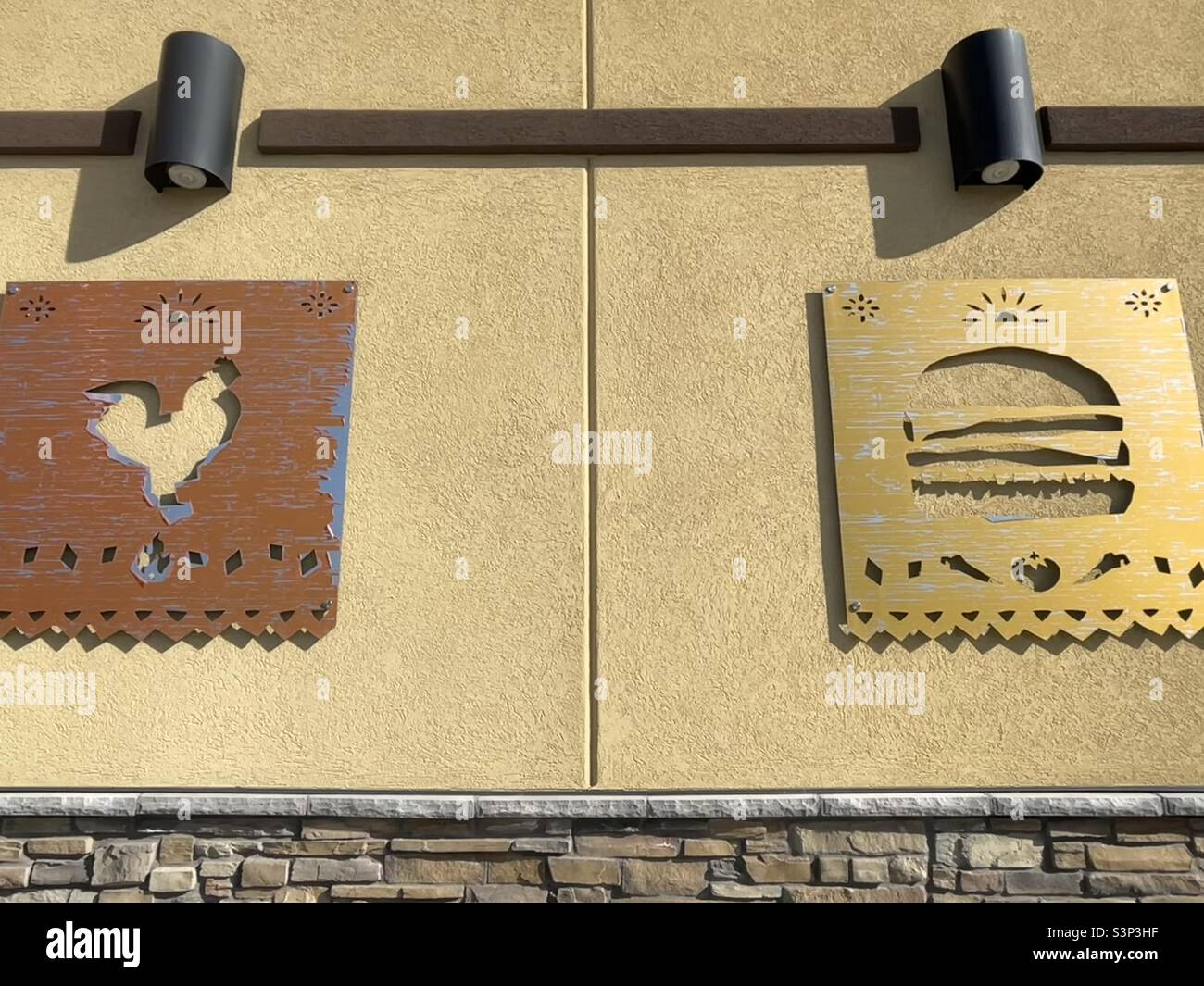 Building side of a local Del Taco fast food restaurant in Utah, USA. Rustic metal cutout signage of a chicken and burger can be seen adjacent to one another under spotlights for nighttime illumination - Smartphone Captured Stock Image
