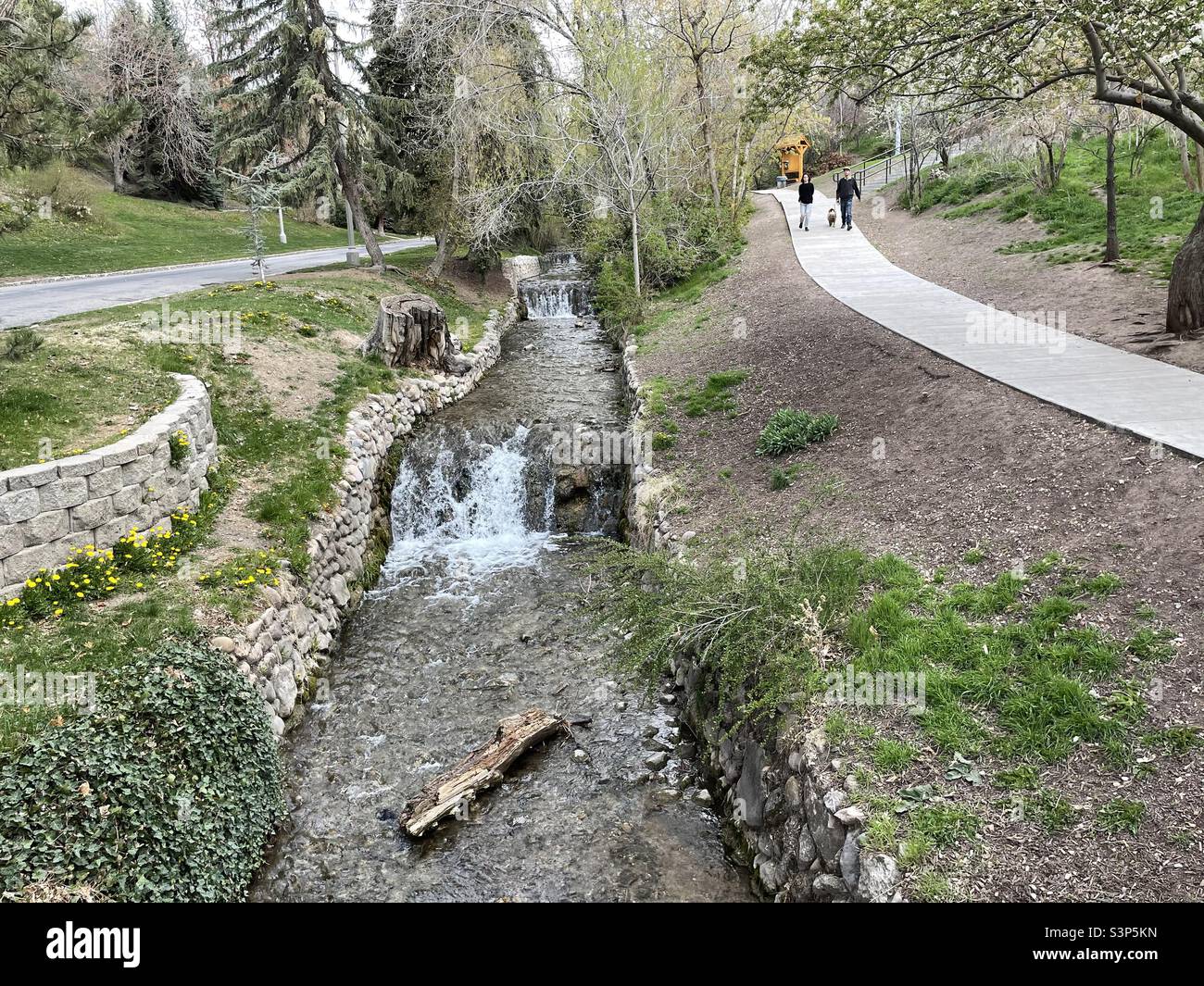 A couple can be seen on the path to the right walking their dog next to a stream that runs through Memory Grove Memorial park in Salt Lake City, Utah, USA on a nice spring day. - Smartphone Captured Stock Image