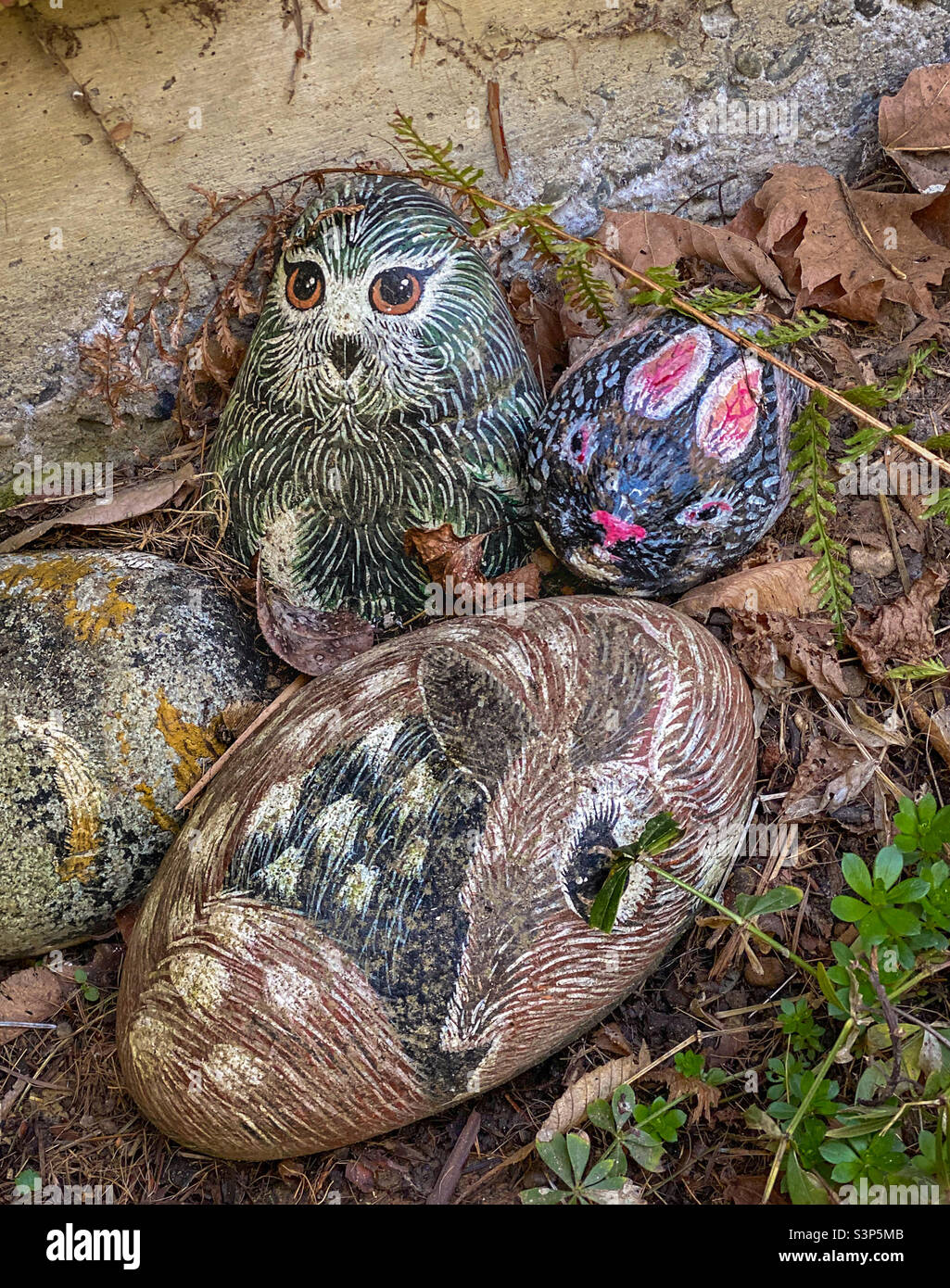 Round painted rocks depicting an owl, rabbit and fawn lie among dried leaves beside the foundation of a house, their bright eyes appearing to stare warily at an approacher. - Smartphone Captured Stock Image