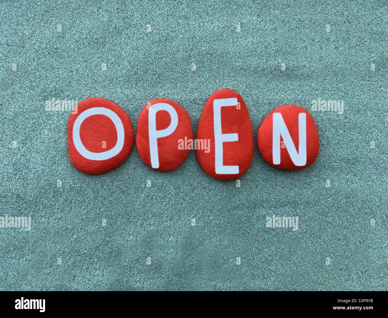 Open word composed with red colored stone letters over green sand Stock Photo