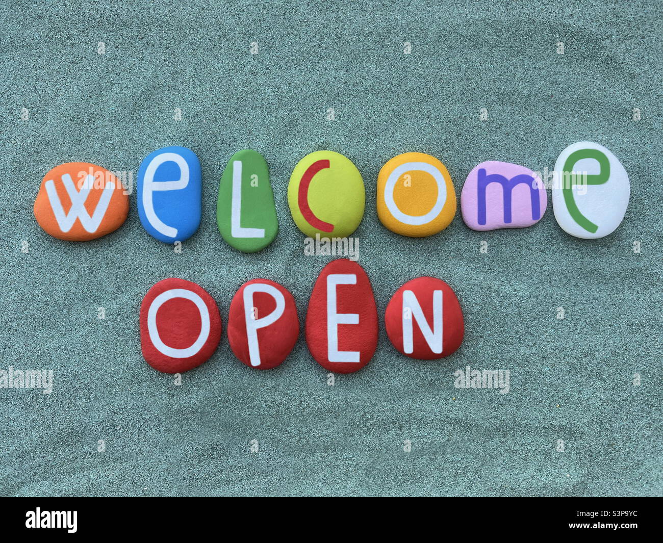 Welcome, Open, creative text composed with multi colored stone letters over green sand Stock Photo