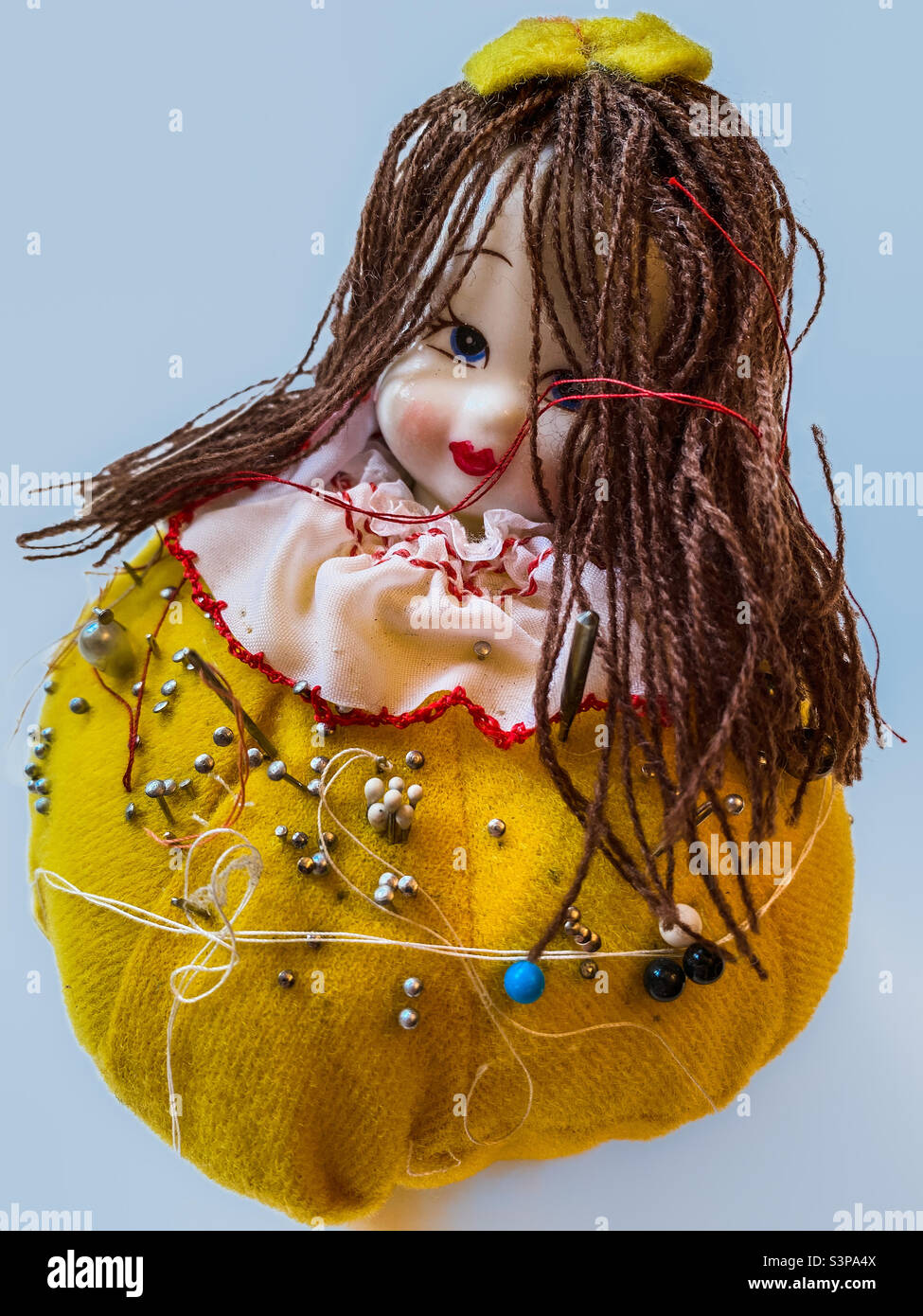 A smiling pincushion doll, made of bright yellow fabric and a ceramic head, looks coquettishly at the viewer from behind her long hair of brown wool (blue background). - Smartphone Captured Stock Image