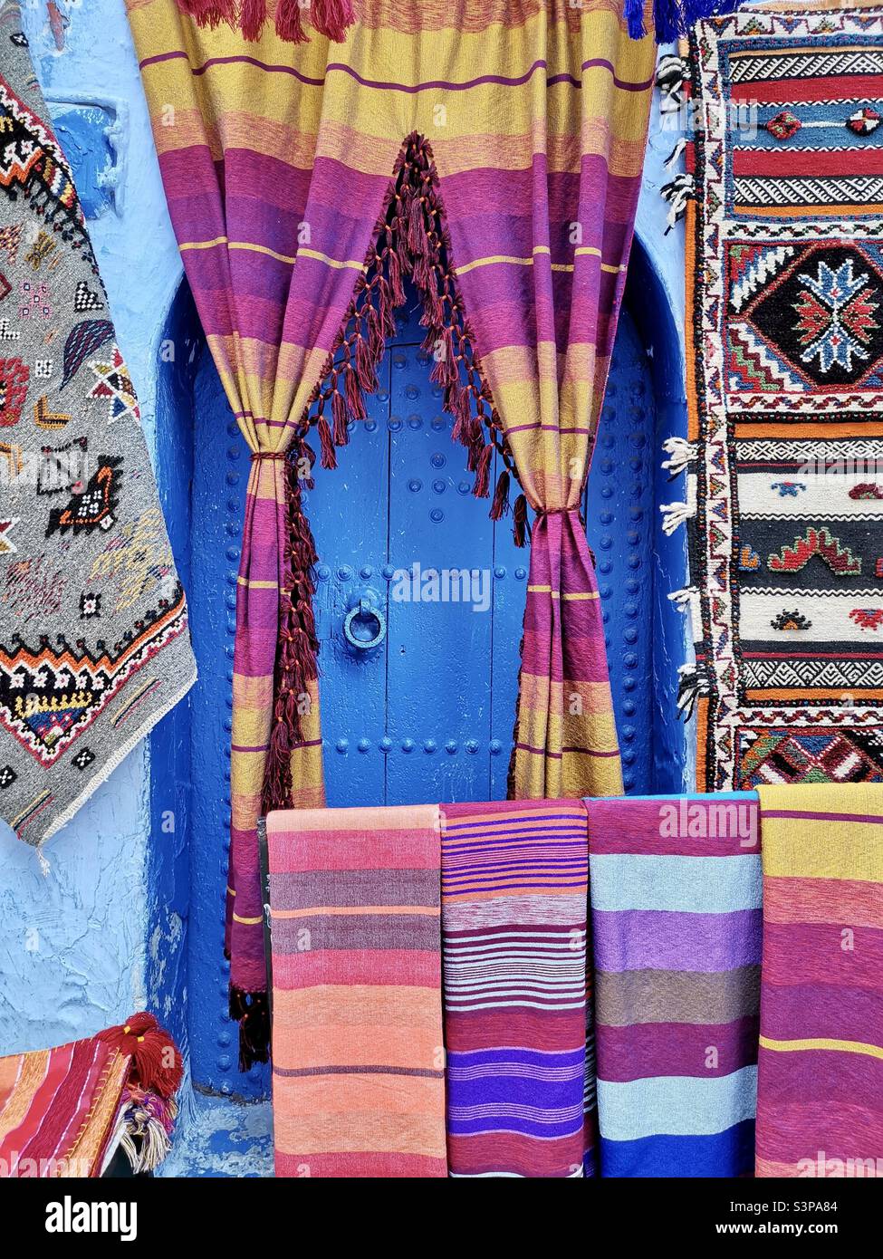 Traditional Moroccan garments ,scarfs and carpets hanged by a blue door in Chefchaouen, Morocco. Stock Photo