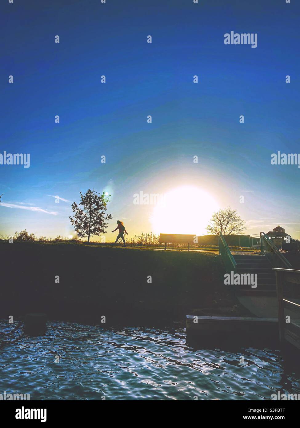 A heavily filtered shot of a scene in a park at sunset.  The sun is large and low, a silhouette of a person walking, a playground in the background  and water from a lake in the foreground. (1) Stock Photo