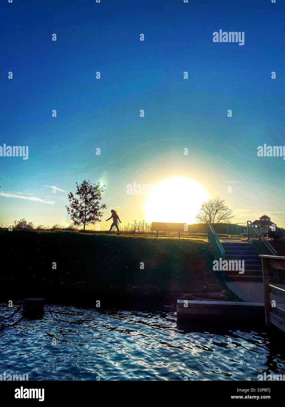 A heavily filtered shot of a scene in a park at sunset.  The sun is large and low, a silhouette of a person walking, a playground in the background  and water from a lake in the foreground. (2) - Smartphone Captured Stock Image