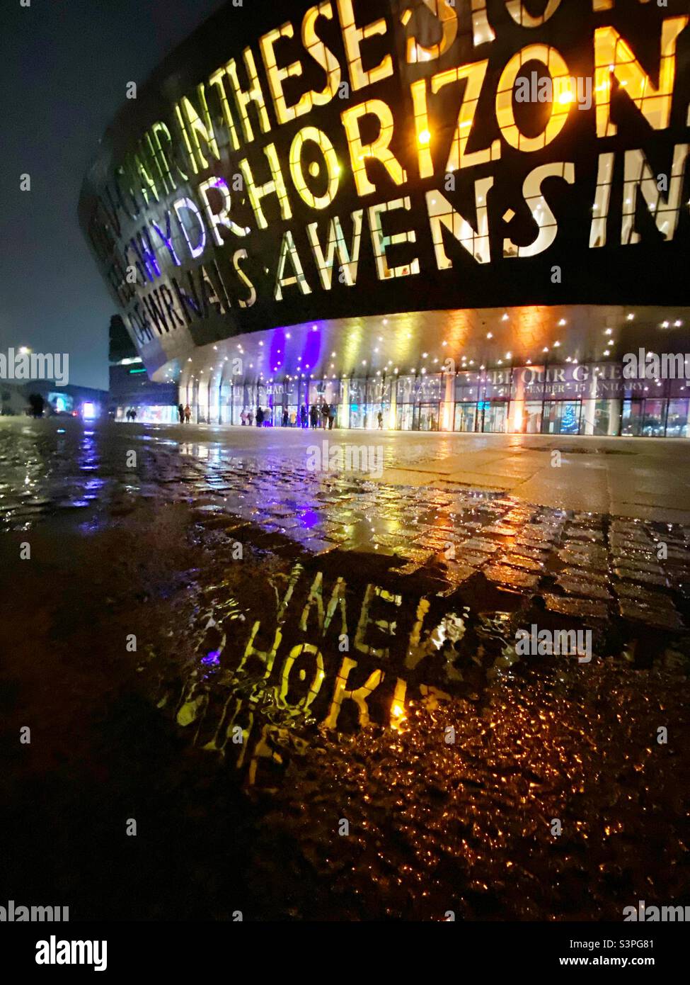 A photograph of the Wales Millennium Centre on a dark raining evening. Reflections of the lights in the puddles. Lit up in Pride colours. Theatre-goers waiting outside Stock Photo