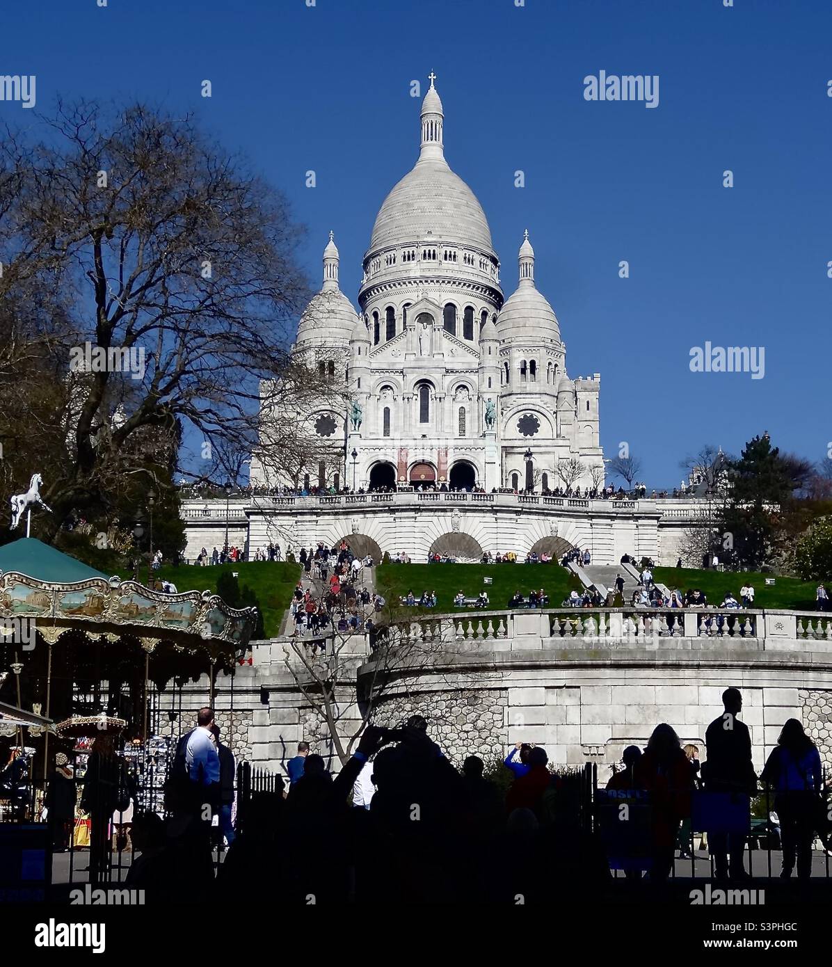 Basilica of Sacre-Coeur in Paris, France, seen from the bass of the butte of Montmartre. It is 131m high (430 feet) and has 5 domes. A children’s carousel and silhouetted figures are seen below. Stock Photo