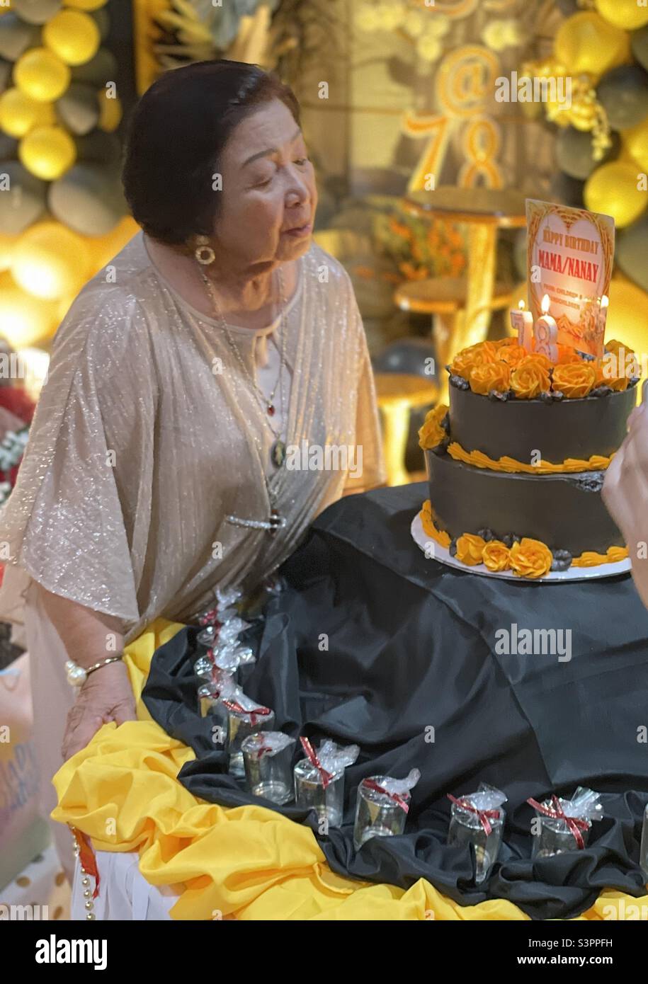 Old asian lady blowing her birthday cake and celebrating 78 years of life in a gold and blavk themed party Stock Photo