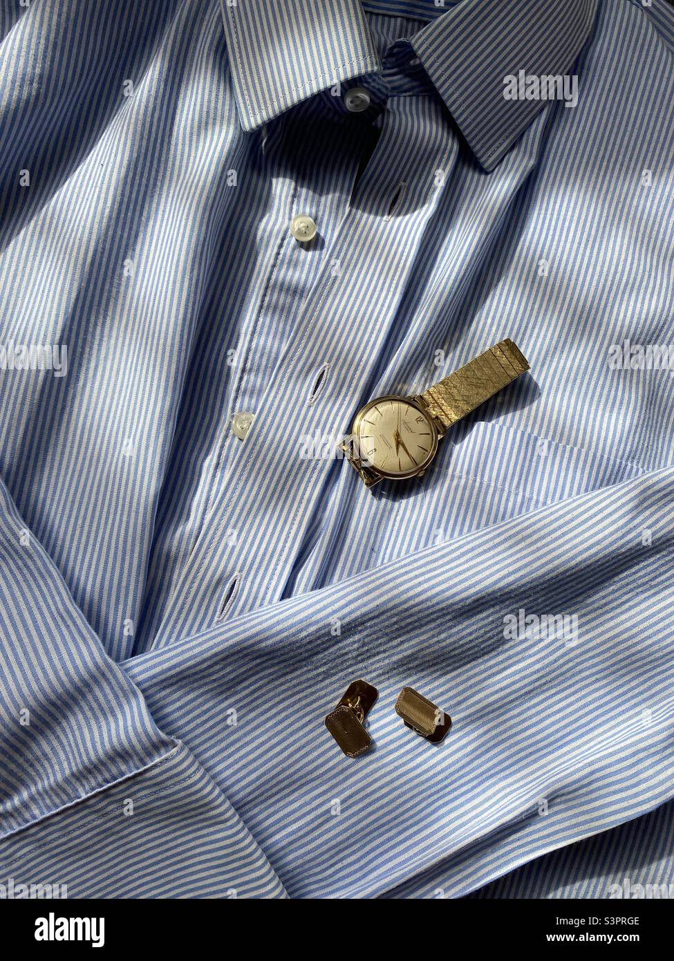 Gentlemen’s elegant blue and white striped shirt with two accessories - a gold wrist watch and gold cuff links - all laid out in the afternoon sun ready to use. Stock Photo