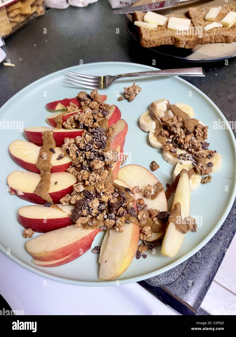 Sliced apple with almond butter drizzle topped with oats and chocolate nuggets on plate with a fork, toast and butter in background Stock Photo