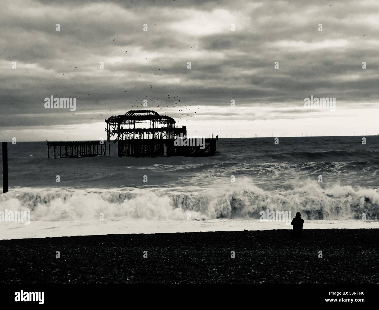The ruined pier at Brighton, England, in stormy seas, with big breaking waves and gales at sunset with a lonely silhouetted figure looking out from the beach with sea birds circling above the ruins. Stock Photo