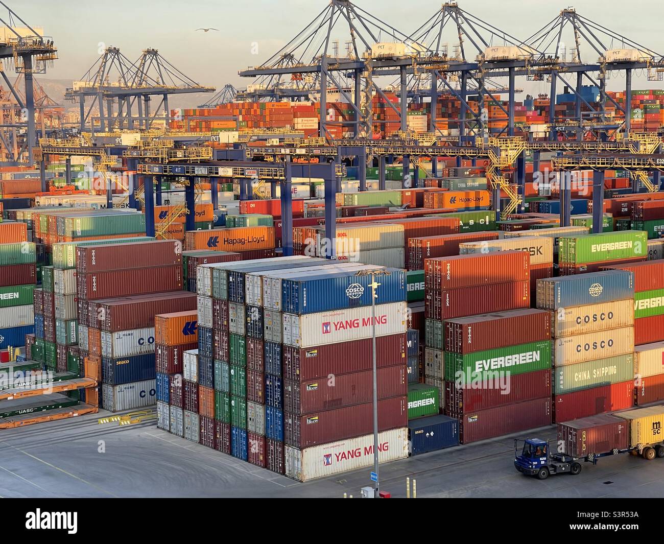 Cargo Container Terminal with plenty of stowed containers from different shippers in port of Piraeus, Greece. In background are gantry cranes and blue sky during sunset. Stock Photo