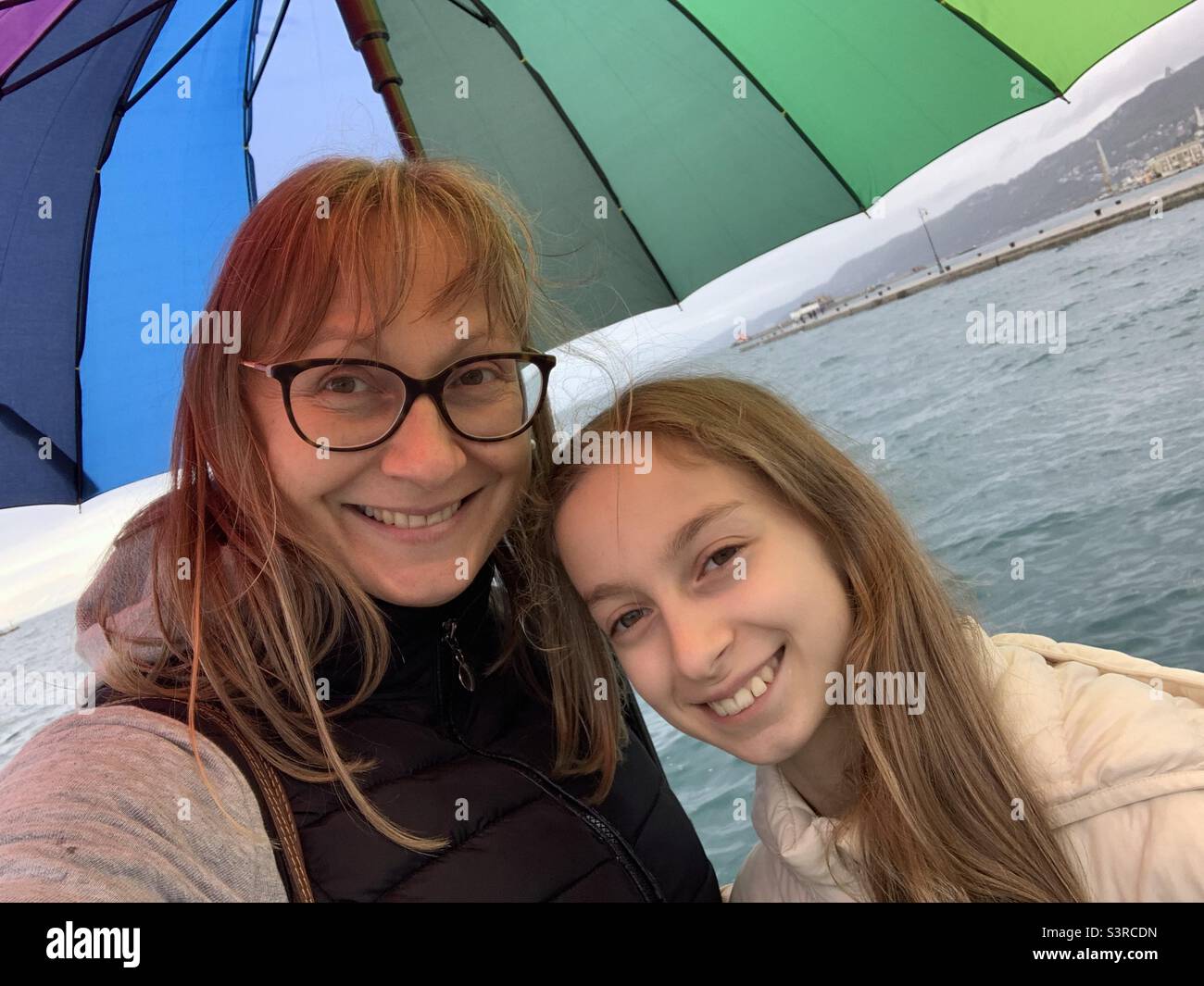 Mother and daughter under rainbow umbrella on a rainy day by the sea ...
