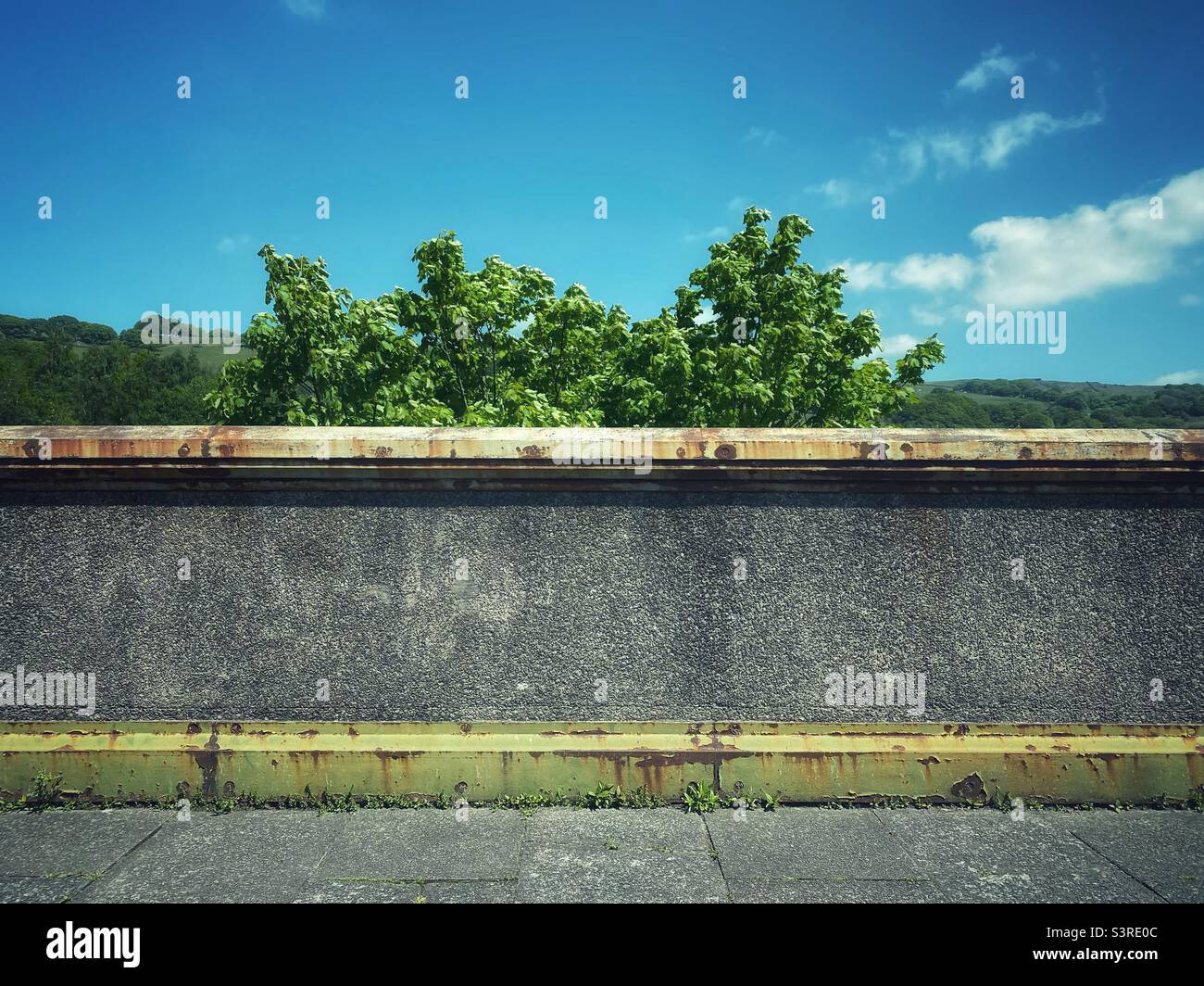 A photograph of a blank wall and rusted metal structure of a bridge, with greenery and sky background. Copy space horizontal. Urban decay scene. - Smartphone Captured Stock Image