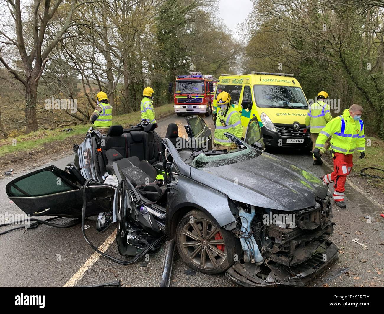 A car which is heavily damaged following a road traffic collision is surrounded by emergency service personnel. Stock Photo