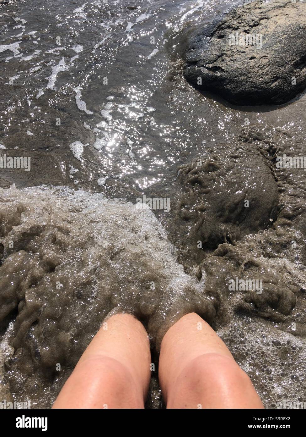 A woman has feet submerged in mud by the sea. Stock Photo