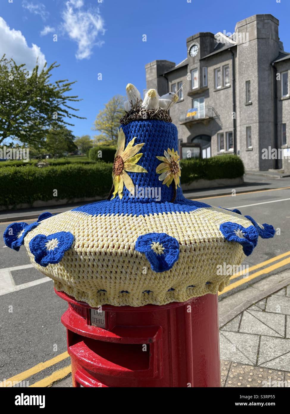 A pillar box adorned with a woolly hat showing support for the Ukraine with its blue and yellow colours, standing in front of the Memorial Hall in Cricceth, North Wales - Smartphone Captured Stock Image