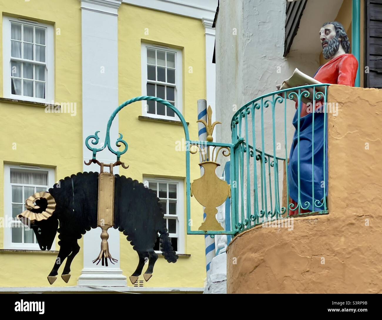 A sculpture of a saintly looking figure seems to read from a balcony overlooking a hanging sign depicting a ram in Portmeirion Village in Wales - Smartphone Captured Stock Image