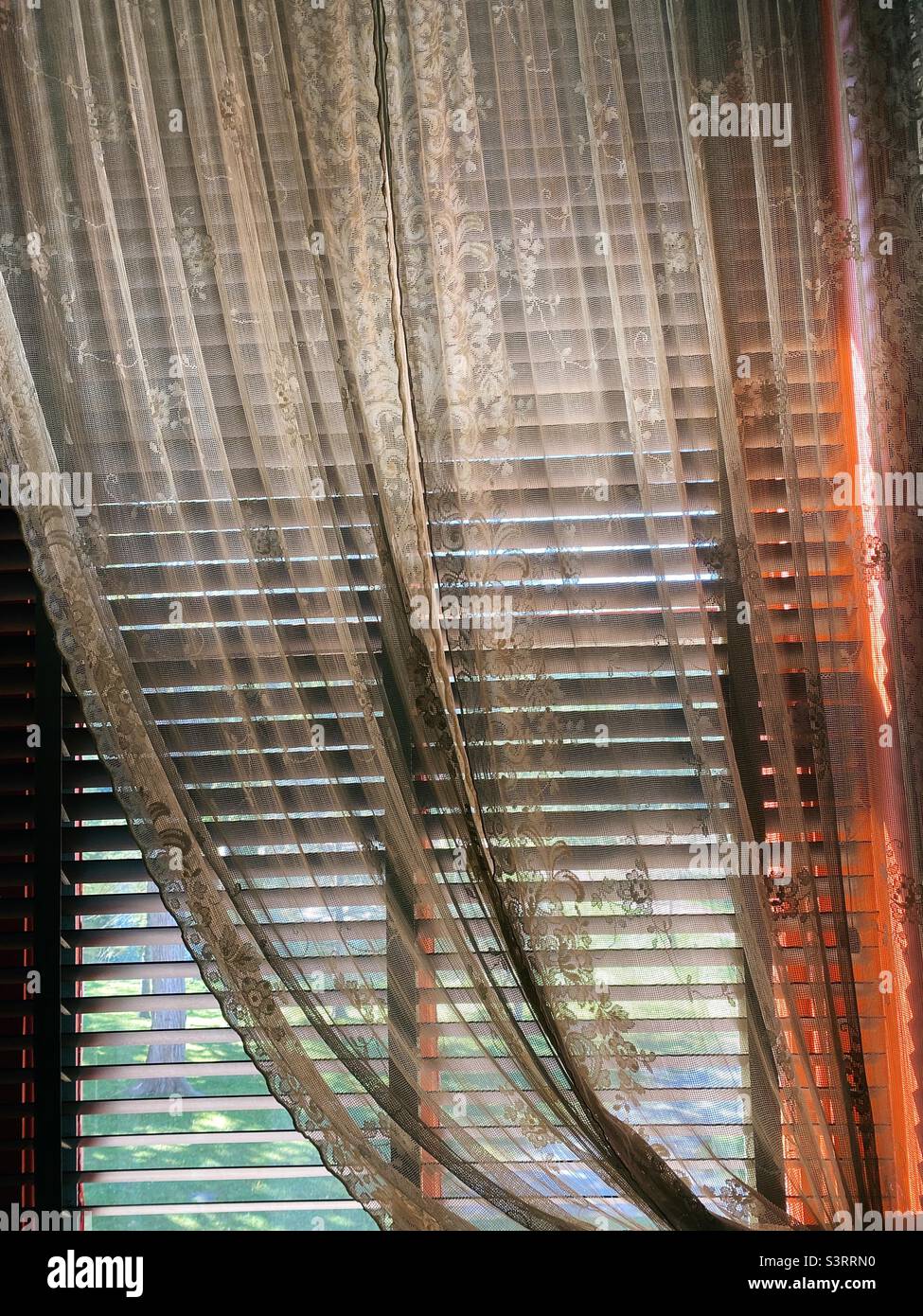 Lace curtains hanging over partially closed blinds with light filtering in. - Smartphone Captured Stock Image