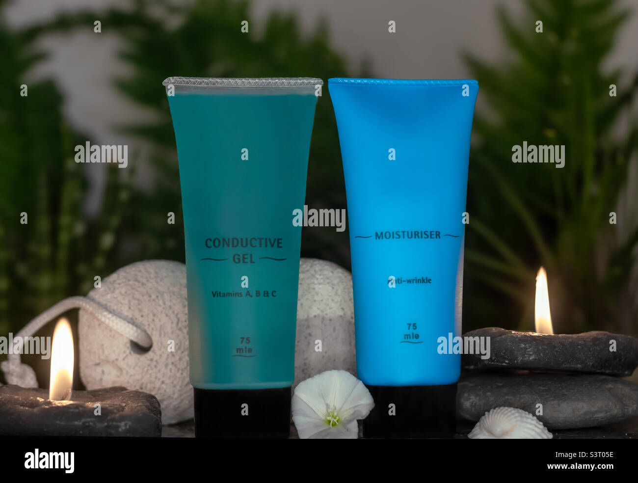 Blue bottles of beauty products displayed with candles, pebbles, pumice and a flower against a green, leafy blurry background - Smartphone Captured Stock Image