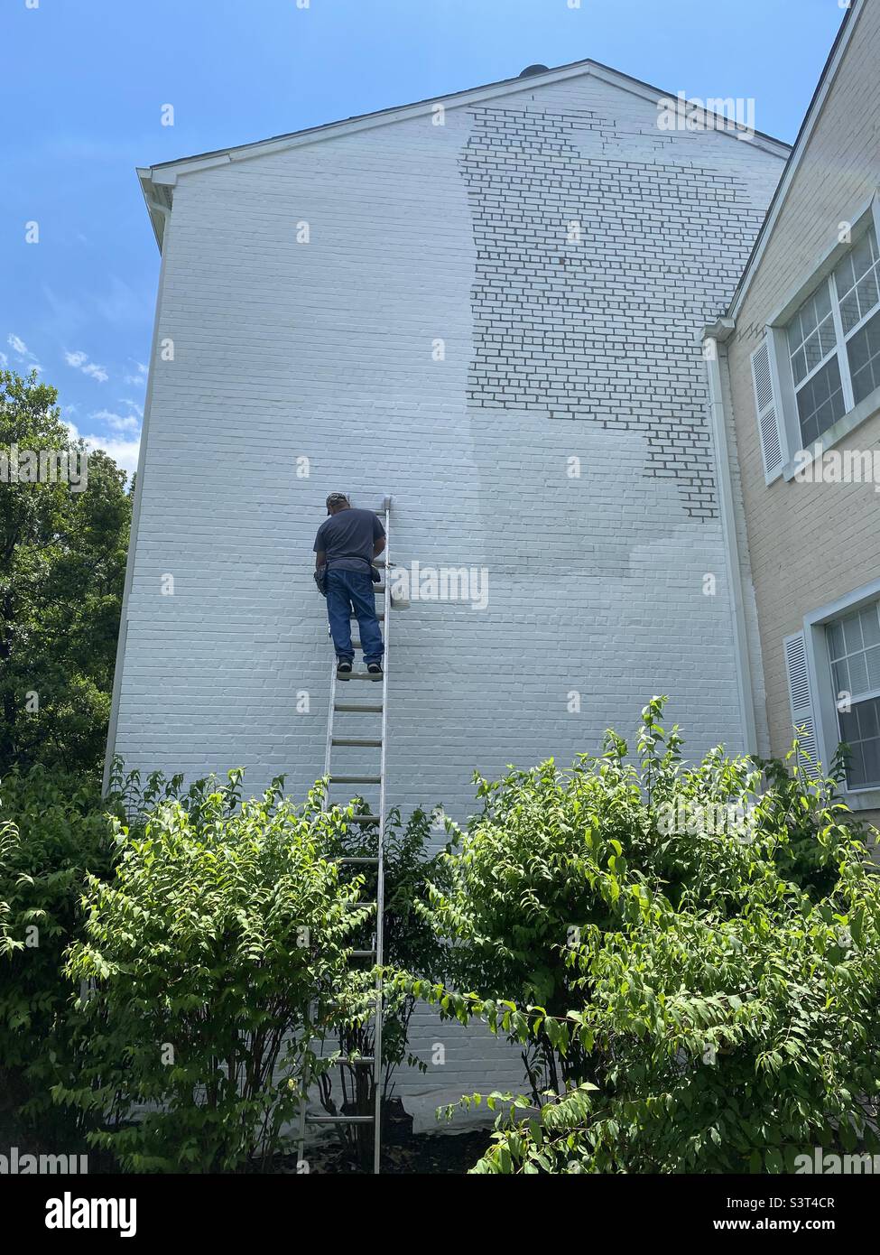 A big job. A man stands on a tall ladder painting the side of a brick building. Stock Photo
