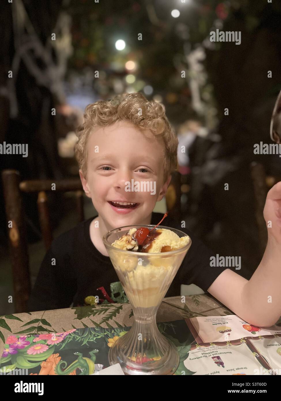 A young 4 year old boy with tight curly blonde hair smiles whilst enjoying  a vanilla ice cream topped with caramel sauce and a Glaze cherry on top at a Cafe in the London Theatre area of England. Stock Photo
