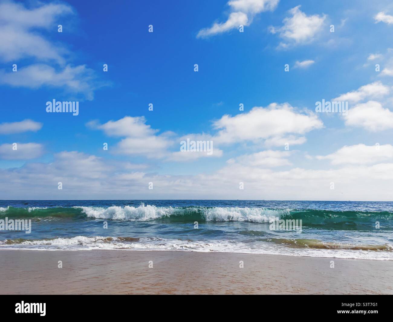 Seascape with waves breaking over sandy beach under a blue sky and white clouds. Stock Photo