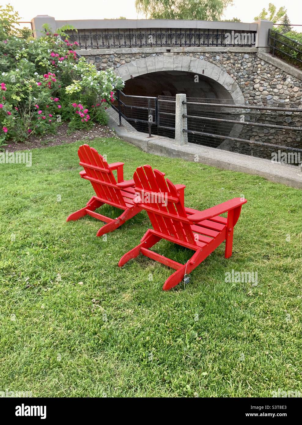 Two bright red Muskoka chairs by a stone bridge over Patterson Creek waiting for guest. - Smartphone Captured Stock Image