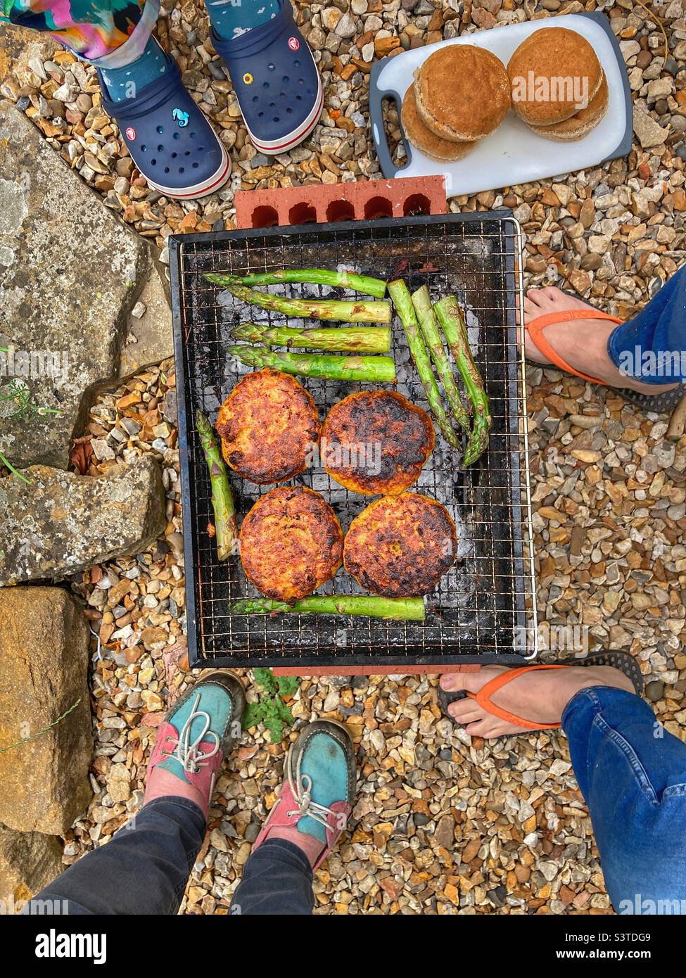 Three people standing around a portable barbecue, cooking chicken katsu burgers and fresh asparagus. High angle view from above Stock Photo