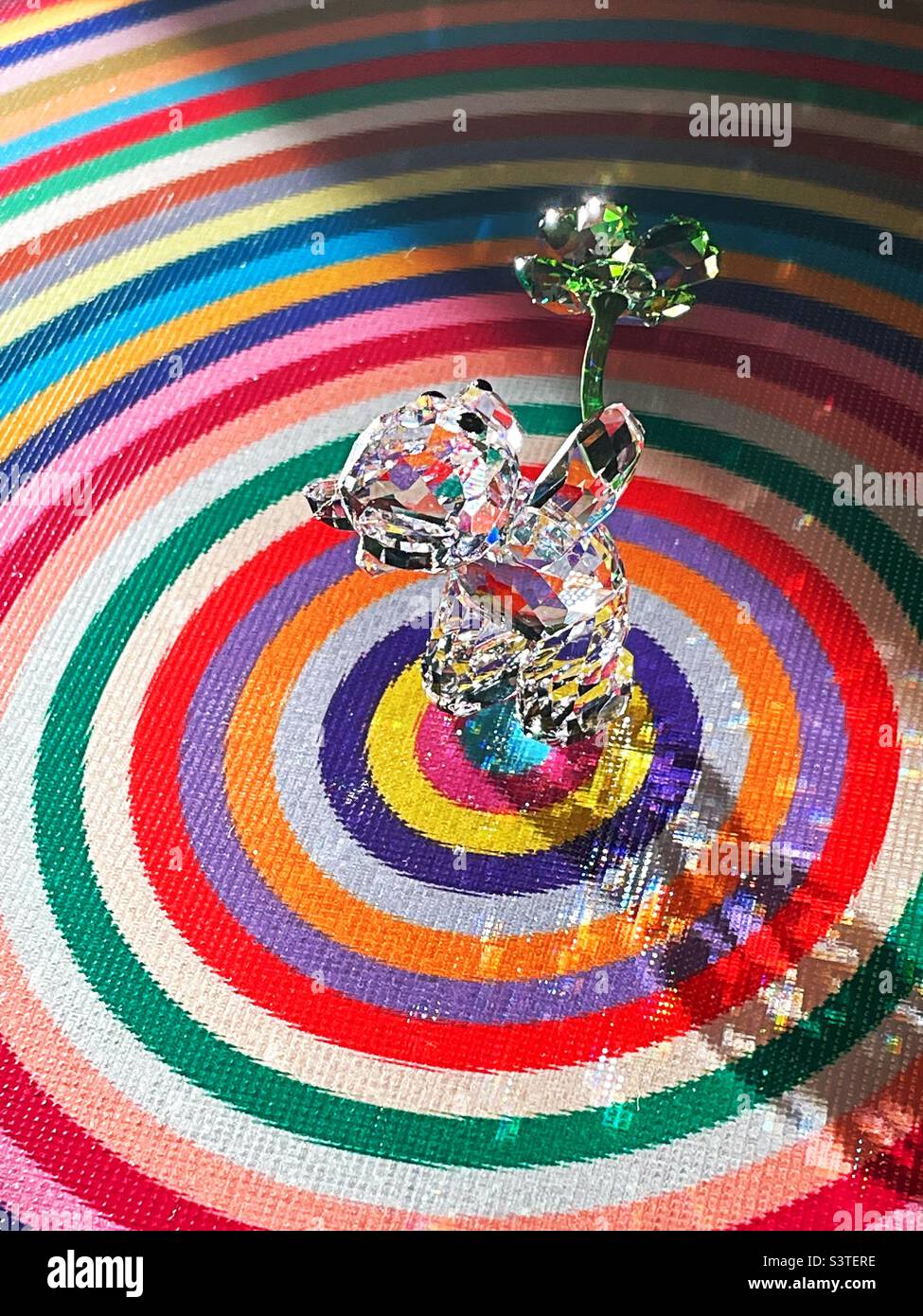 Crystal figurine of a bear cub playing with a shamrock on a colorful circular cutting board with bright sunlight, 2022, USA - Smartphone Captured Stock Image