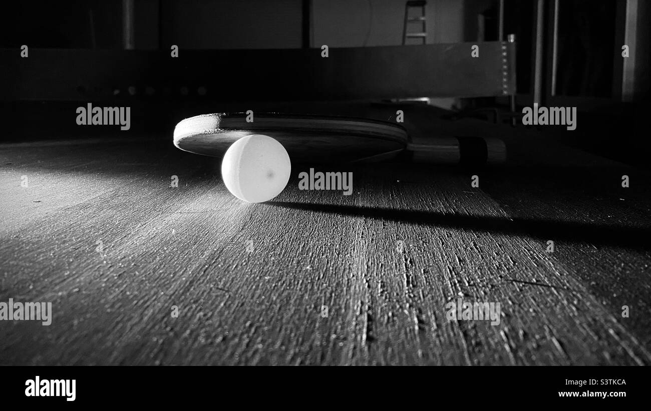 Shallow focus black and white view with dramatic lighting of a ping pong or table tennis ball and bat resting on a worn table - Smartphone Captured Stock Image