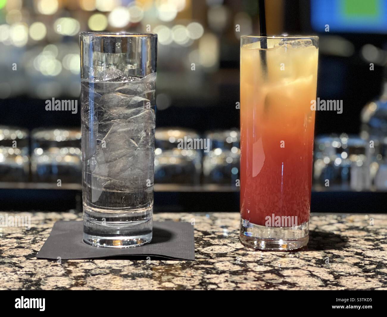 Tall glasses on bar top, one containing ice and water, the other filled with tequila sunrise - Smartphone Captured Stock Image
