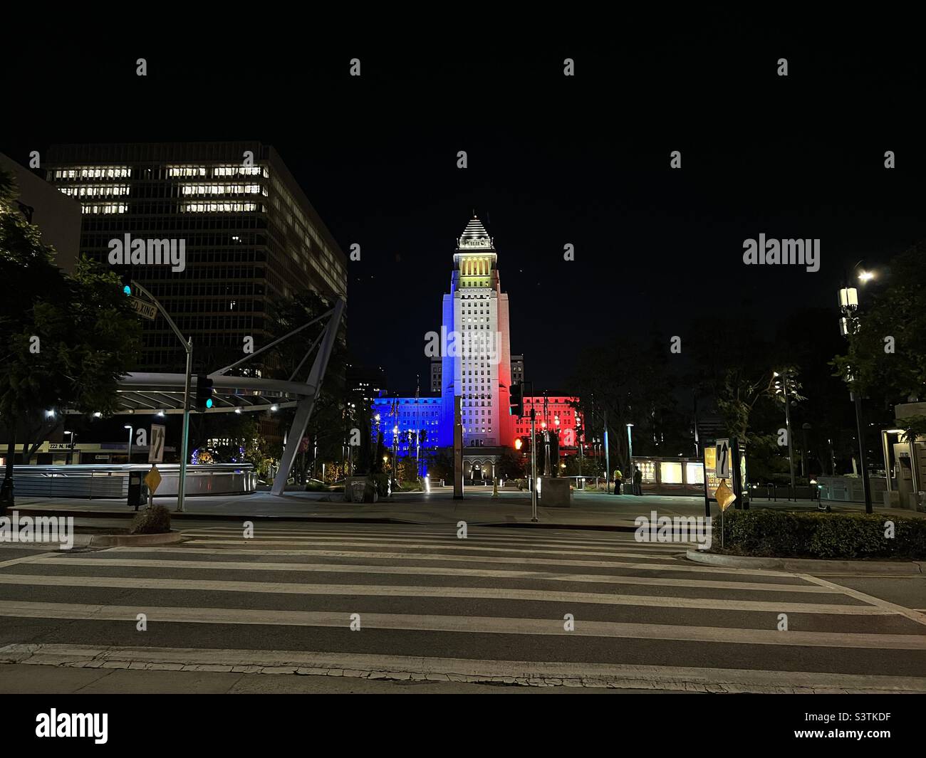 LOS ANGELES, CA, OCT 2021: wide view of City Hall in Downtown at night, with blue, white and red lighting on the building. Pedestrian crossing and LA Metro subway station in foreground - Smartphone Captured Stock Image