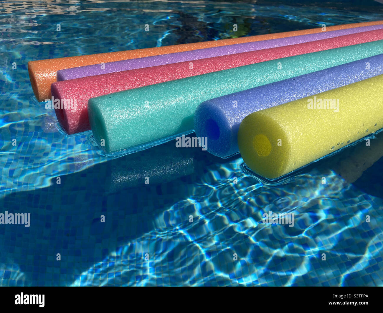 Rainbow coloured pool noodles floating in sparkling water of swimming pool. Stock Photo