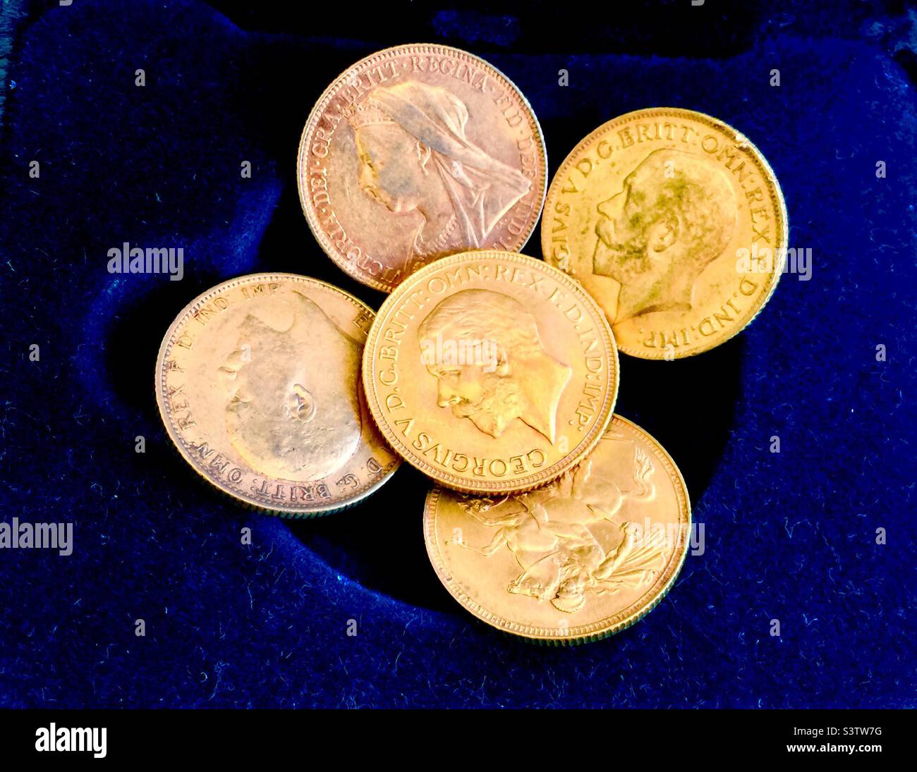 Five antique gold sovereign coins arranged on a rich blue velvet backdrop. Stock Photo