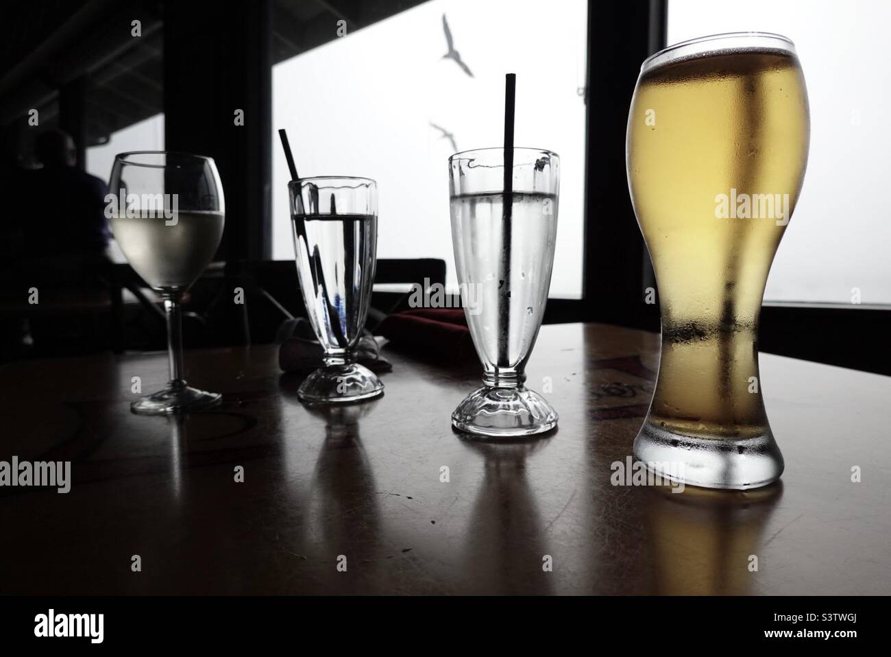Silhouetted white wine, water glasses and beer, with focus on nearest glass at a seaside bar with pelicans flying outside the window - Smartphone Captured Stock Image