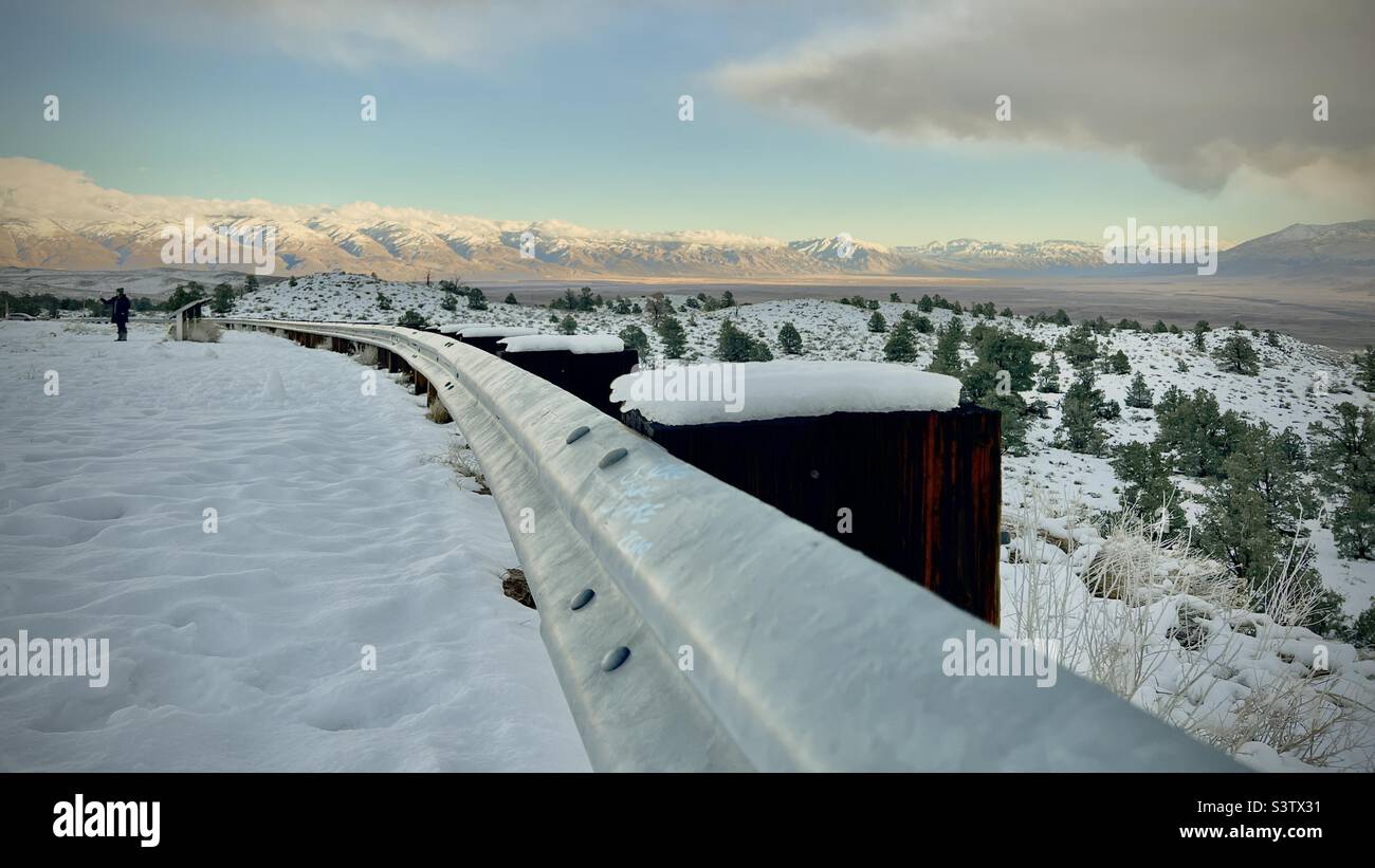 Looking along roadside safety rail, towards snow-covered mountains in the distance, near Bishop, California. Dark storm clouds overhead and over mountains - Smartphone Captured Stock Image