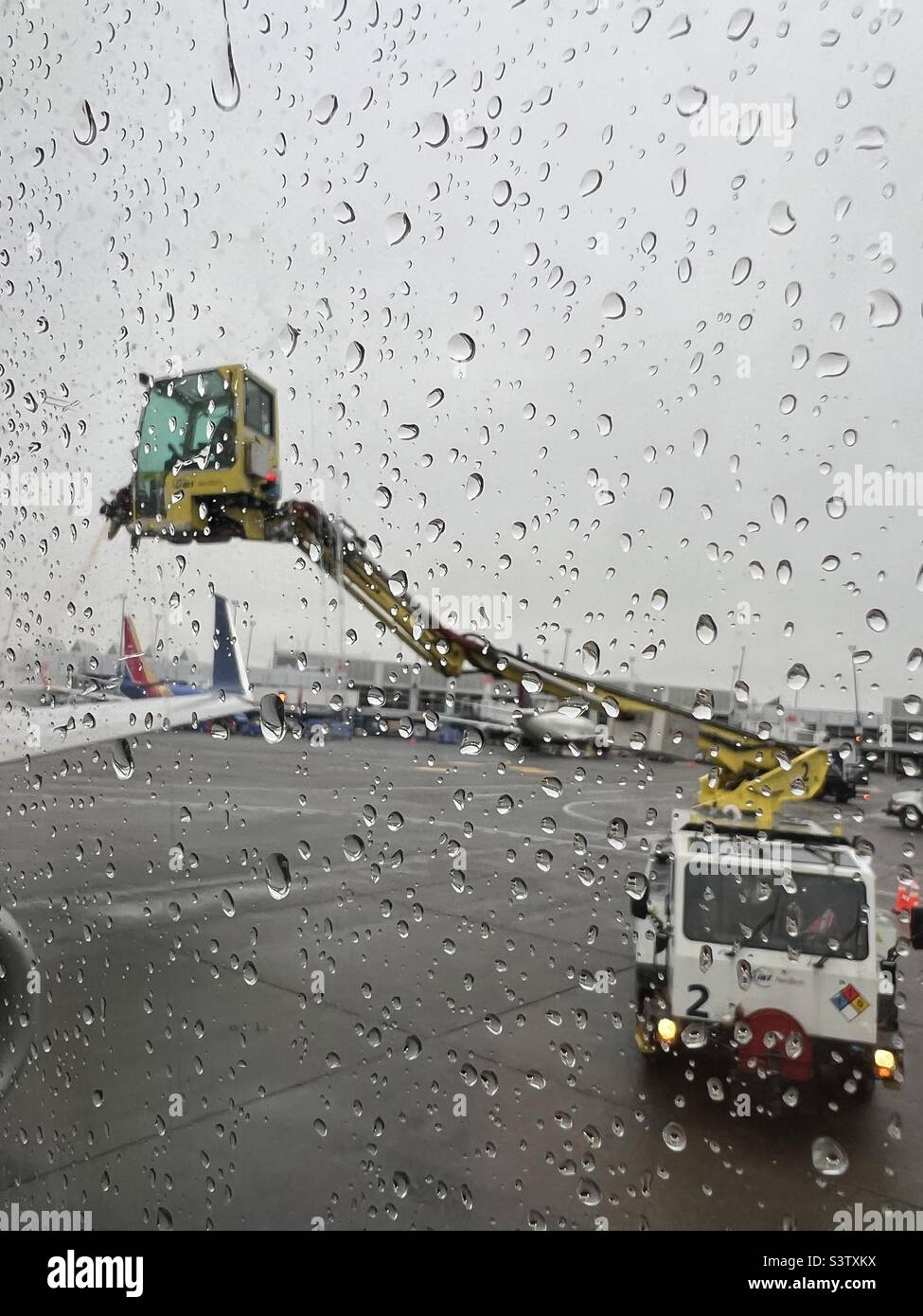 Vertical view, with focus on rain drops on passenger jet's window as workers, in soft focus, spray de-icing fluid on wings using specialized equipment in background - Smartphone Captured Stock Image
