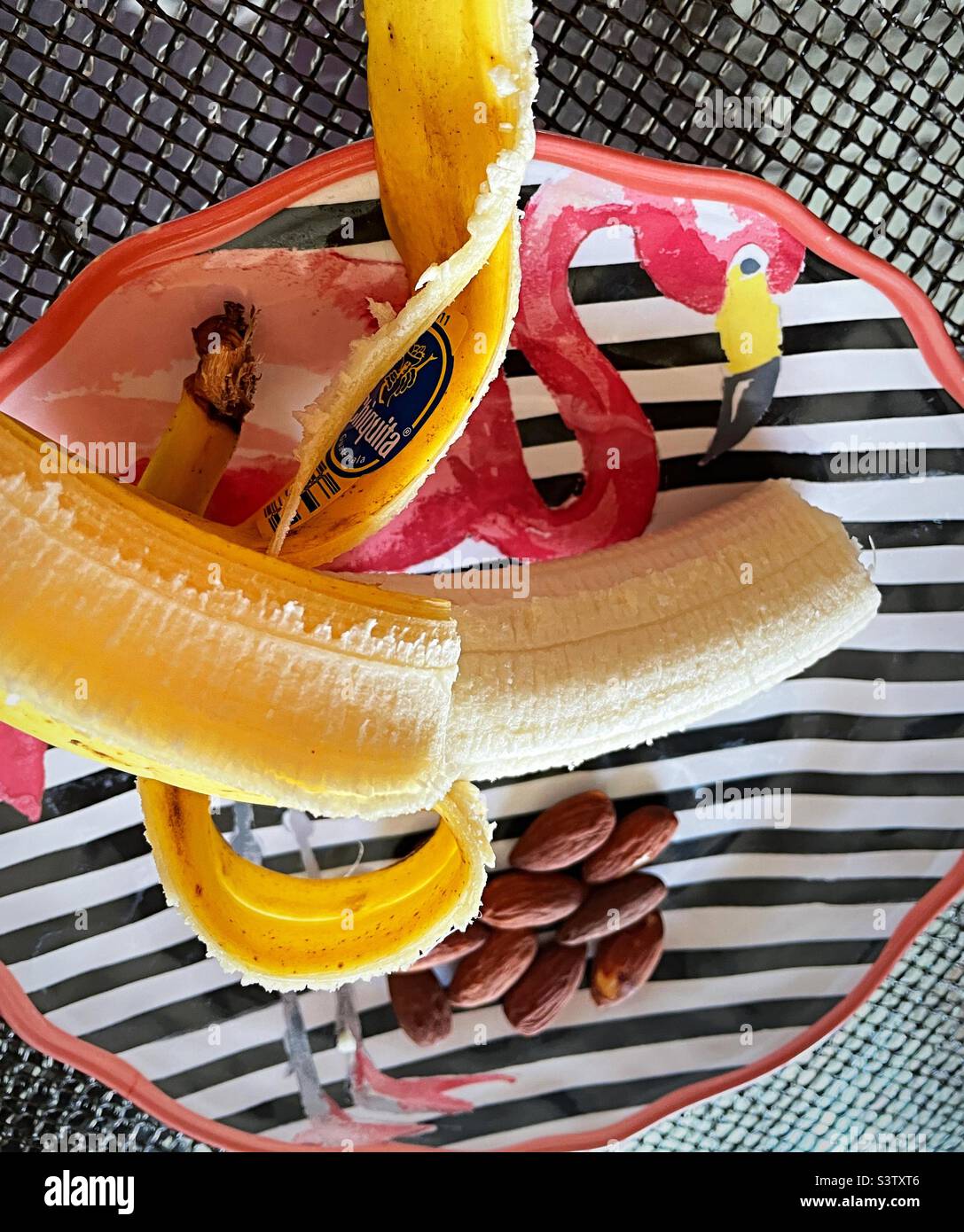 Flat lay of an afternoon snack of a half eaten banana and almonds on a striped flamingo plate, 2022, USA Stock Photo