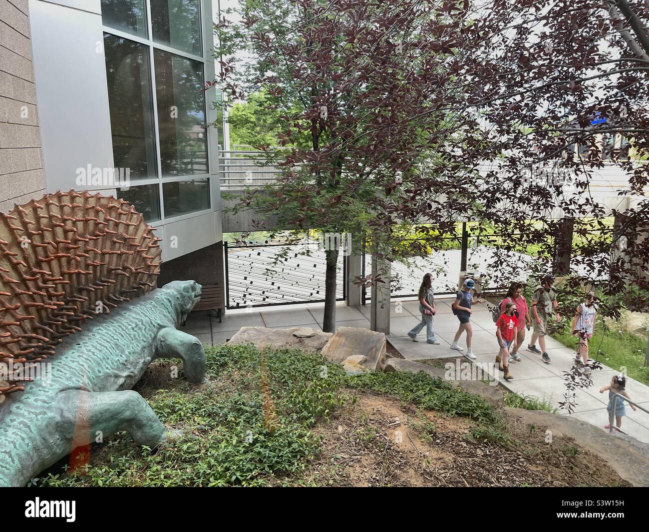 Outdoor grounds at the Dinosaur Museum in Vernal, Utah, USA. Just another day here as visitors and tourists casually walk past yet another skulking dinosaur in the landscaping. - Smartphone Captured Stock Image
