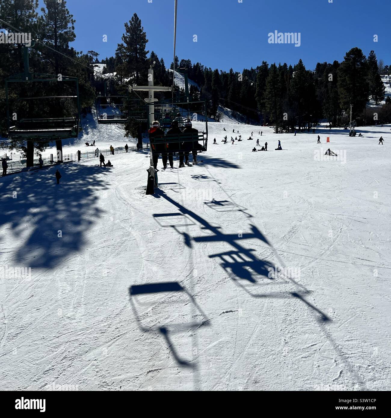 Anonymous riders on lift at Snow Summit in California, casting large shadows on snow covered slopes below, with other skiers and snowboarders on the mountain - Smartphone Captured Stock Image