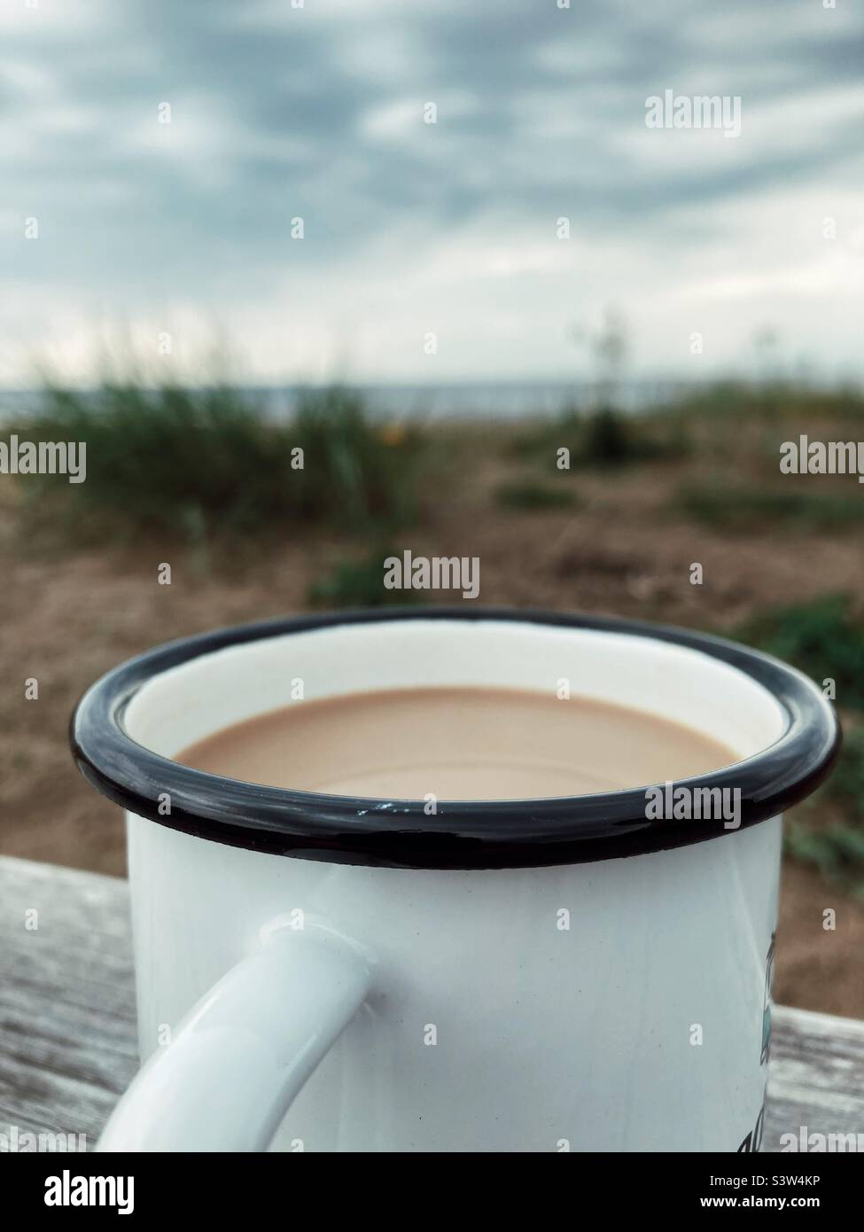 A coffee with milk in a White and Black enamel cup on a Wooden plank at the beach on the West Coast of Sweden Stock Photo