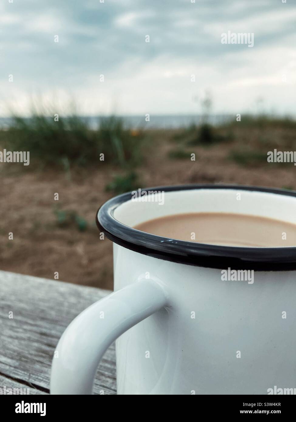A coffee with milk in a White and Black enamel cup on a Wooden plank at the beach on the West Coast of Sweden - Smartphone Captured Stock Image