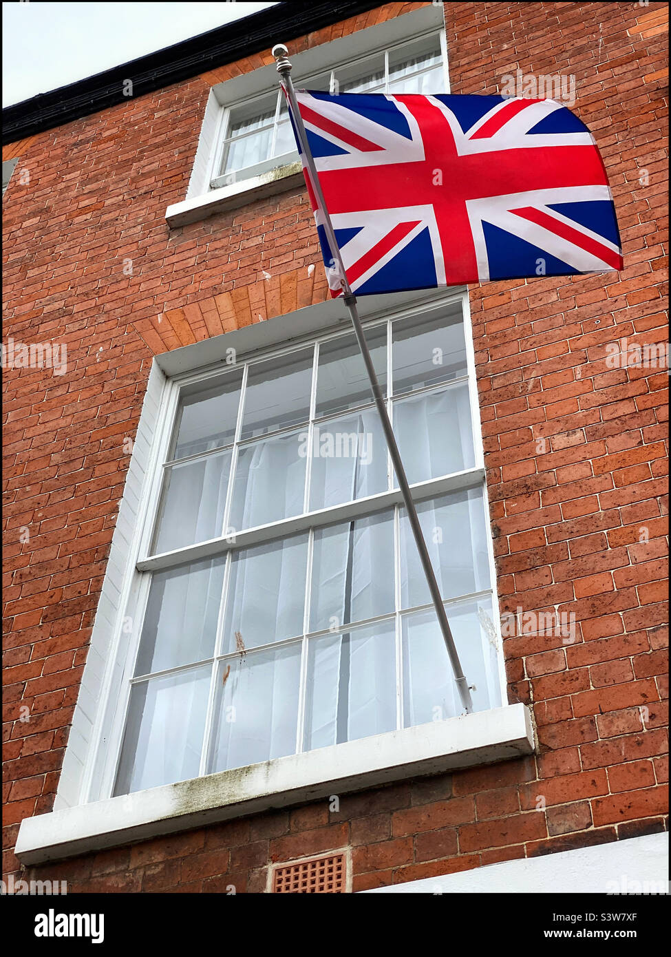 The flag of the United Kingdom (The Union Jack) flutters outside the window of a house somewhere in Great Britain. Photo ©️ COLIN HOSKINS. Stock Photo