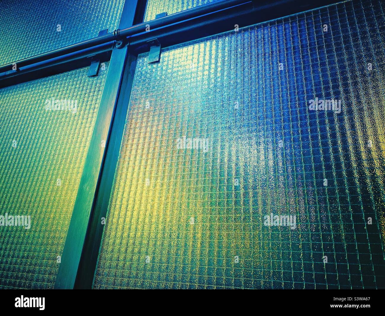 A photograph of industrial interior glass with texture and metal fixings. Blue green light behind. Copy space. Industrial background. Stock Photo