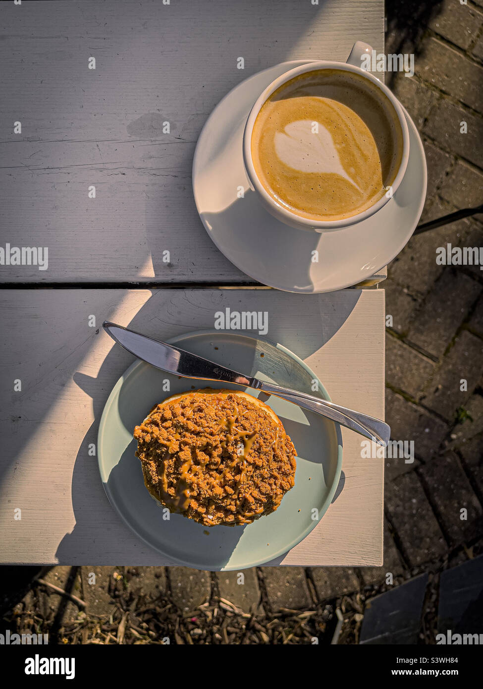 Cup of coffee and a plate with a sticky toffee-topped doughnut on the edge of a white garden table. - Smartphone Captured Stock Image