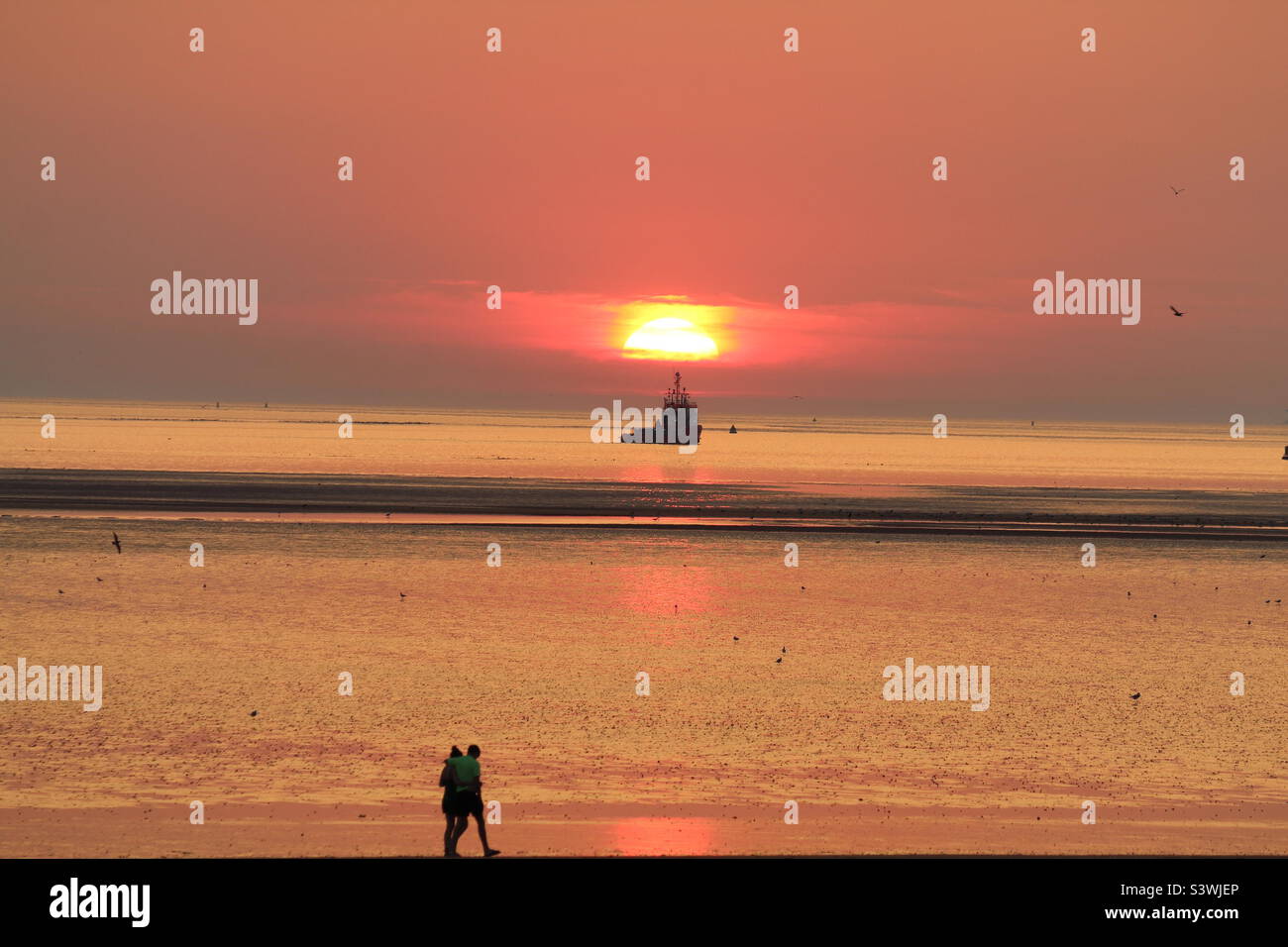 A beautiful sunset at the Beach. A boat can be seen directly under the falling red sun and a couple in love walking. This photo was taken during the exceptionally hot weather and a heatwave. - Smartphone Captured Stock Image