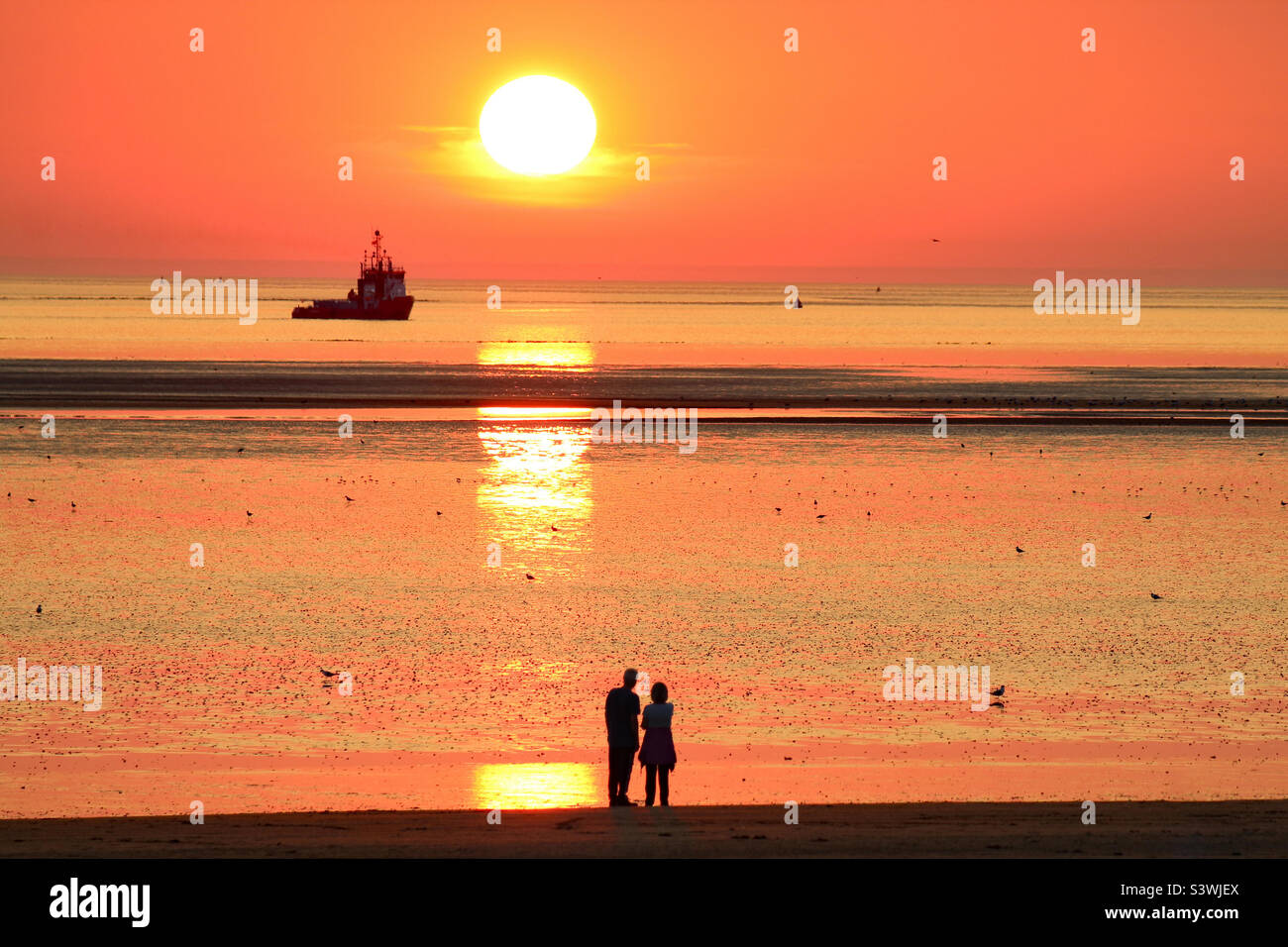 A beautiful sunset at the Beach. A boat can be seen directly under the falling red sun, a couple are gazing at the rare set. This photo was taken during the exceptionally hot weather and a heatwave. - Smartphone Captured Stock Image