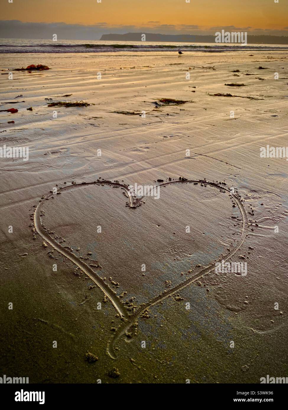 Love on the beach… a hand drawn heart lies on the golden sand of a Southern California beach as the sun sets on the horizon. - Smartphone Captured Stock Image