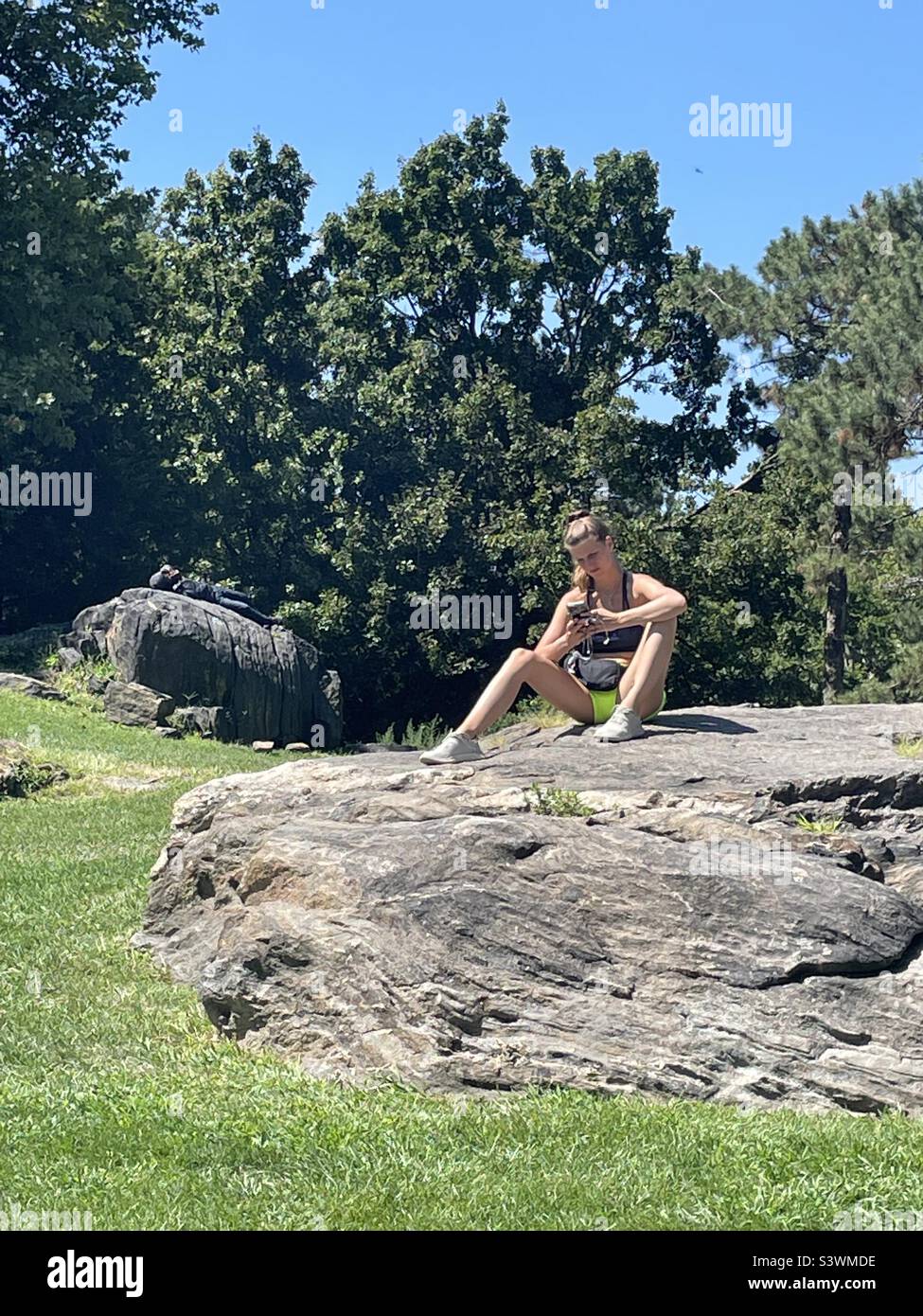 A young woman sits on a rock looking at her cell phone on a summer day in Central Park, New York City, USA - Smartphone Captured Stock Image